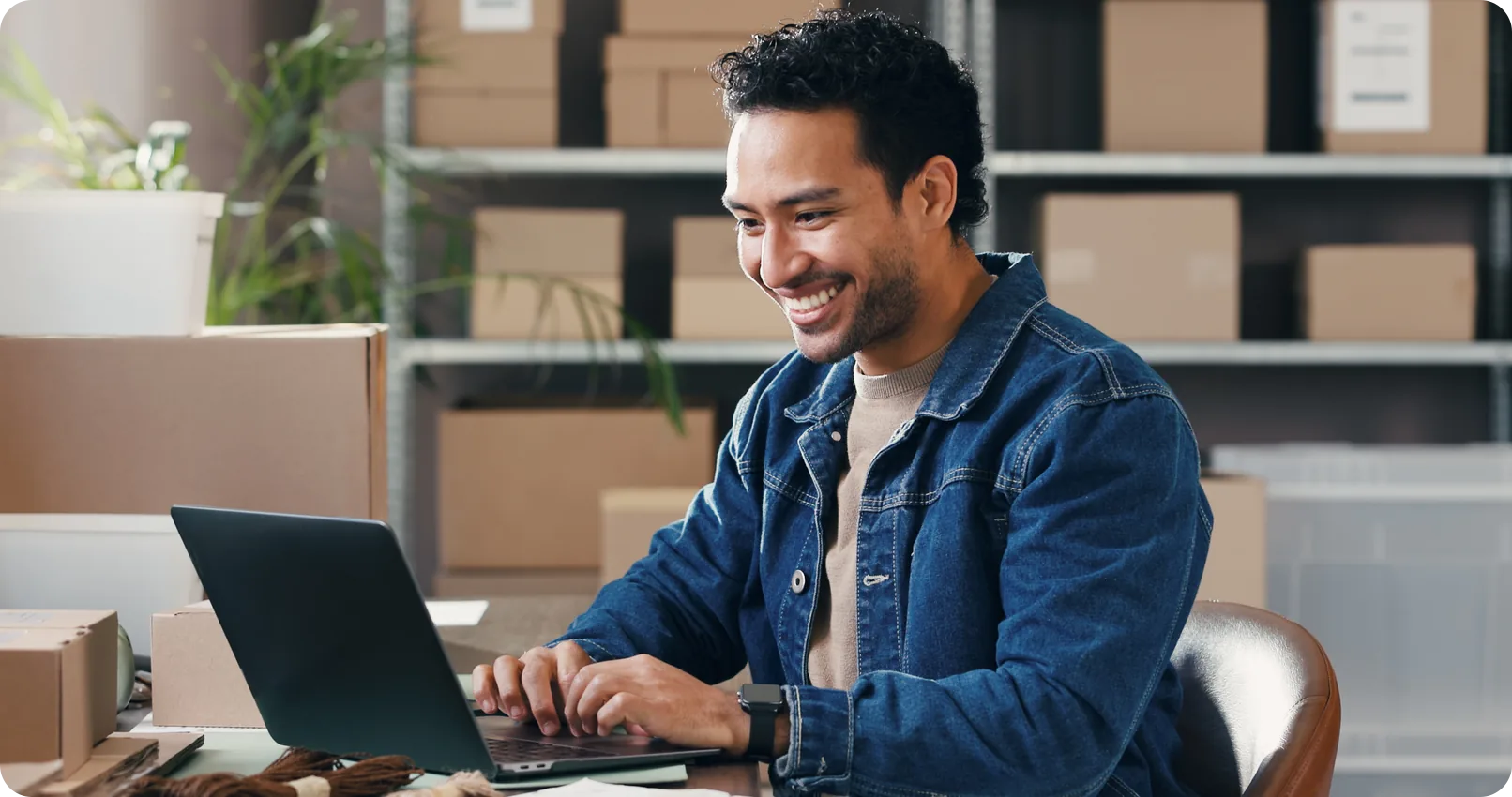 A small business owner smiling while working on a laptop in a stockroom filled with shipping boxes